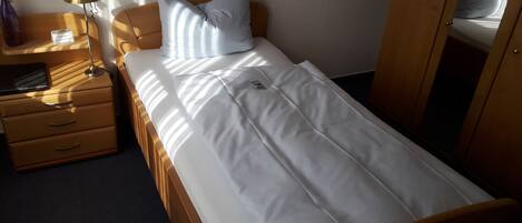Una cama individual con ropa de cama blanca, un cabecero de madera, una mesita de noche con una lámpara y una pequeña mesa con un jarrón de flores.