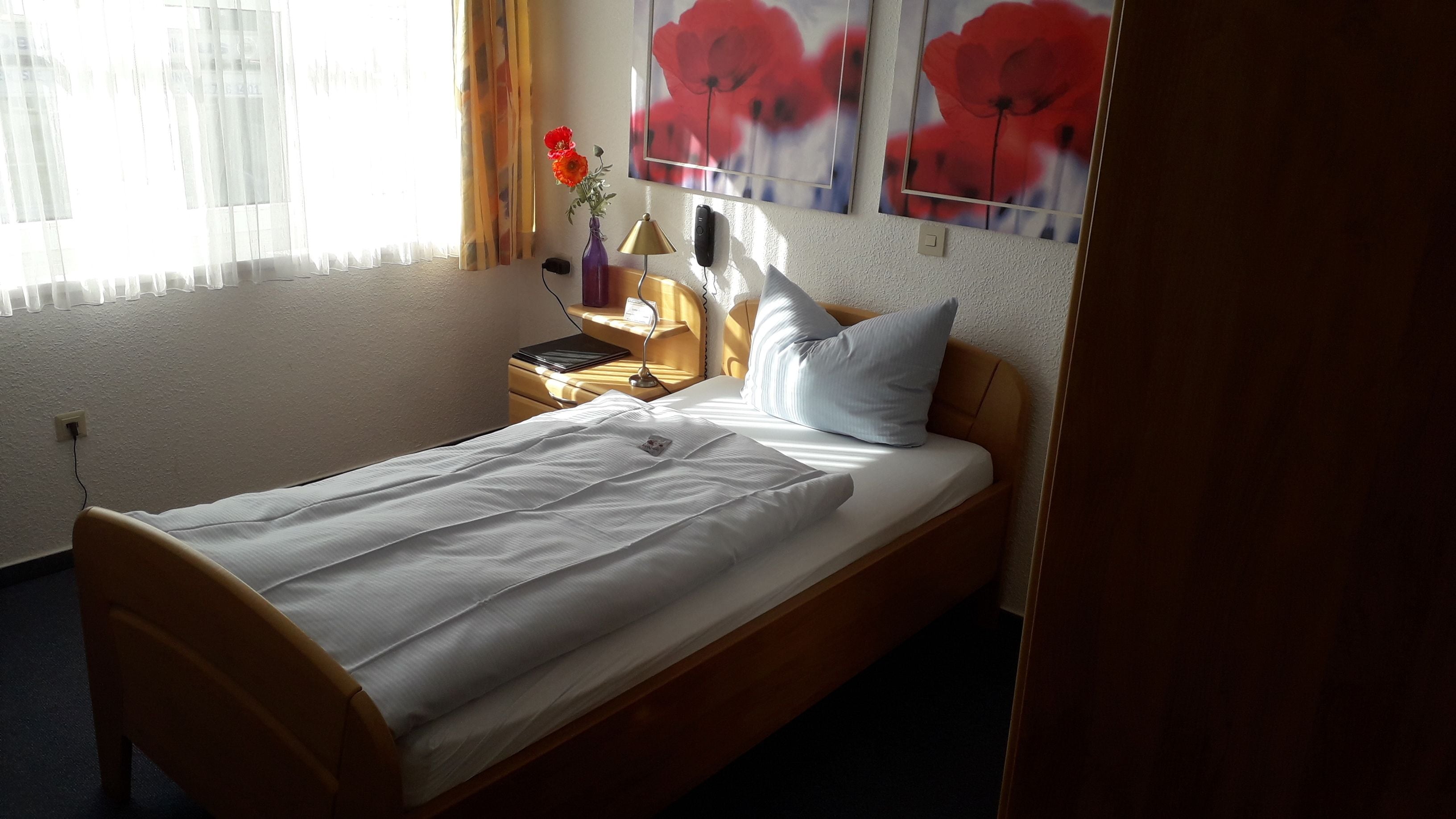 A neatly made bed with white linens, a bedside lamp, a vase with flowers, and a framed picture of red flowers on the wall.