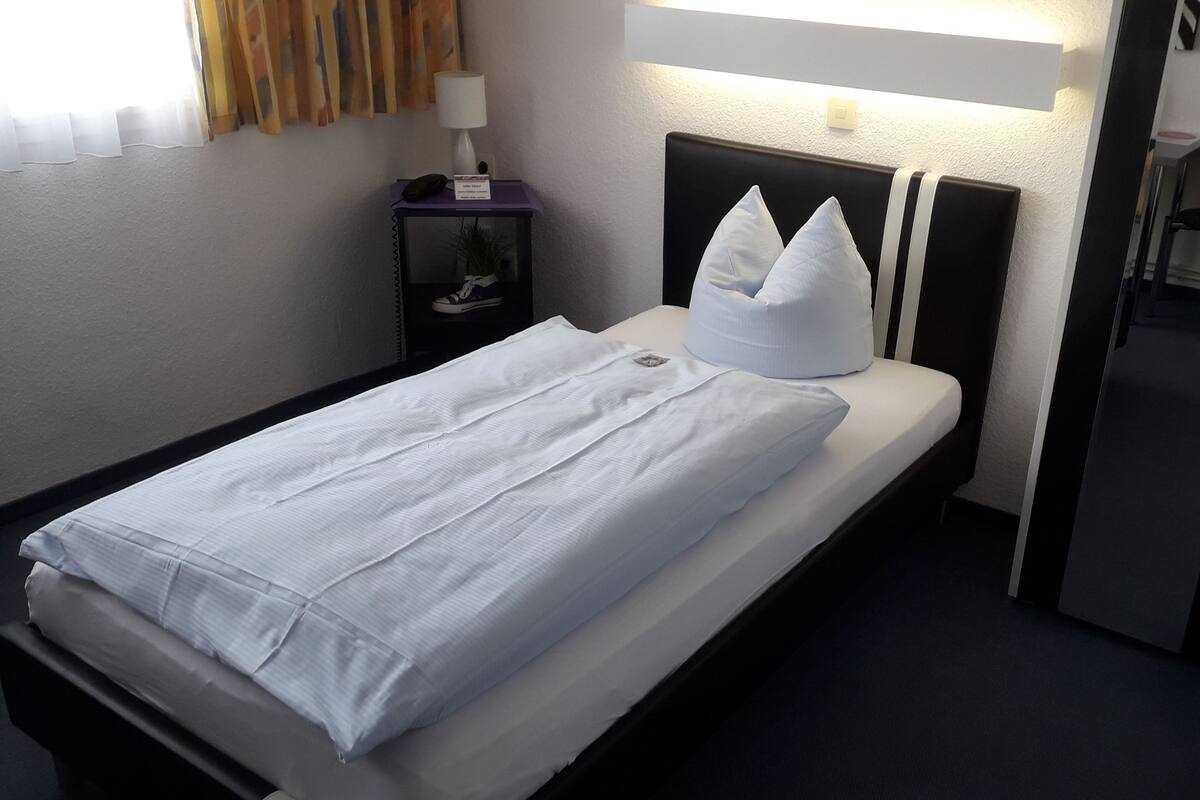 Una cama individual con ropa blanca de cama, cabecera con dos almohadas, mesita de noche con lámpara y una planta pequeña.