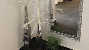 Un pasillo con un espejo grande, paredes blancas y suelo alfombrado. Hay dos plantas en macetas y un par de zapatos en el piso.