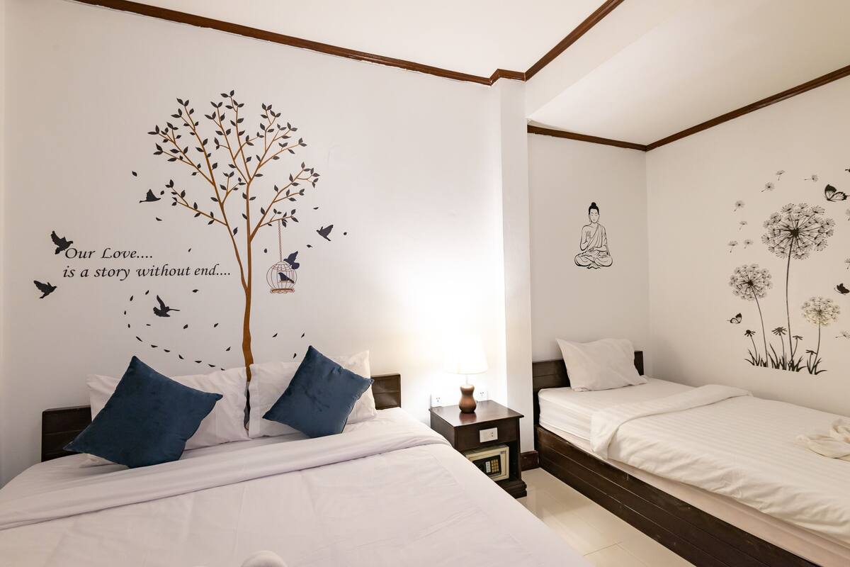 A hotel room with two beds, a nightstand with a lamp, and wall art depicting a tree and a meditating figure.