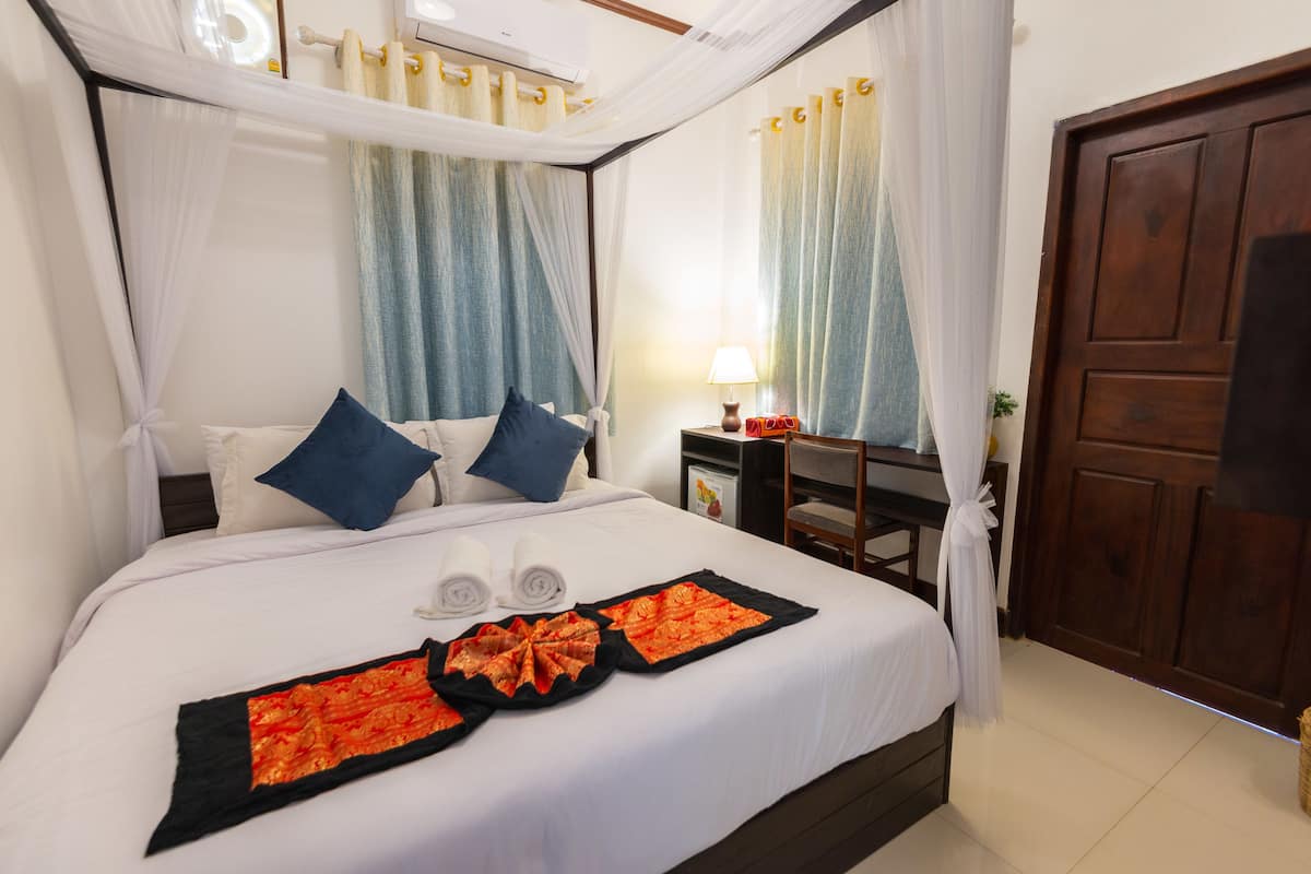 A neatly made bed with a canopy, two pillows, a bedside table with a lamp, and a wooden door.