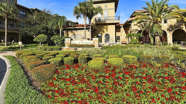 A hotel with a central courtyard featuring manicured hedges and red flowers, a stone facade, and a sign reading "Bella Piazza."
