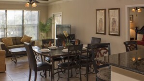 A dining area with a dark wooden table and chairs, a sofa, and a TV. There are framed pictures on the wall and a plant on the table.