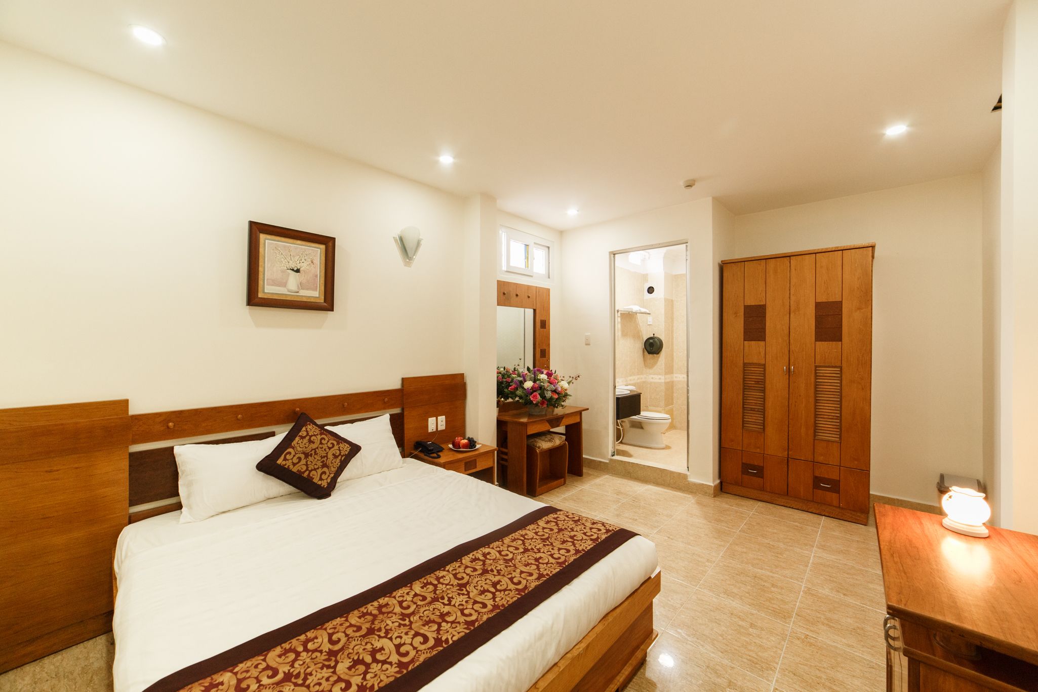 Una habitación de hotel con una cama grande, cabecero de madera, una mesita de noche, un armario y un baño visible a través de una puerta abierta.