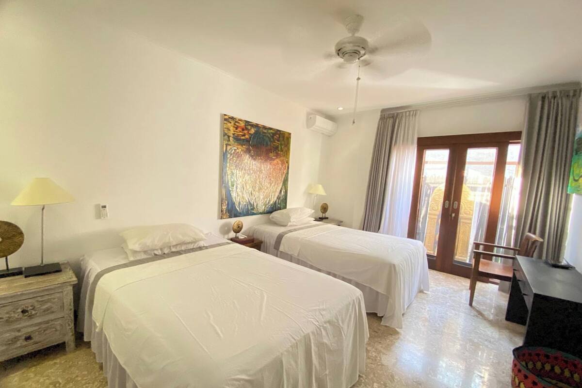 A hotel room with two single beds, a ceiling fan, a painting on the wall, a desk with a chair, and a window with curtains.