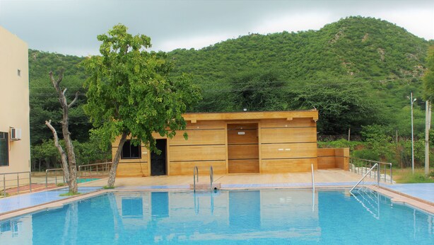 A swimming pool with wooden steps leading into it, surrounded by a building with a simple design and a view of a green hill in the background.