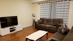 Ein Wohnzimmer mit einem Flachbildfernseher auf einem weißen Standfuß, einem braunen Sofa, einem Holztisch und einem gestreiften Vorhang.
