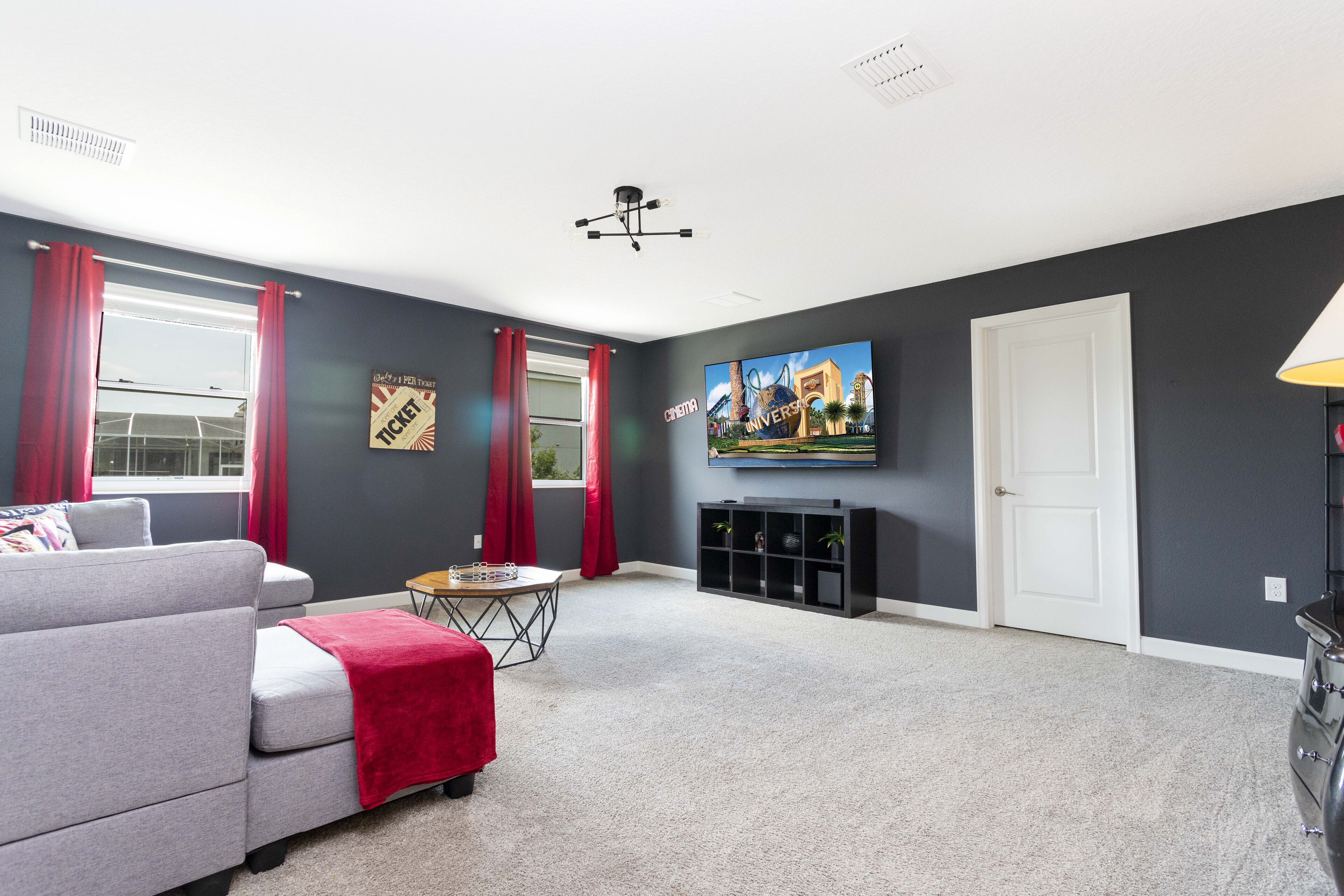 A living room with a grey sofa, red curtains, a flat-screen TV, a ceiling fan, and a small table with a red blanket.