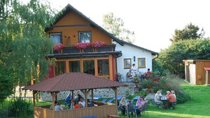 A wooden gazebo with benches and a table, surrounded by a grassy area with a building in the background.