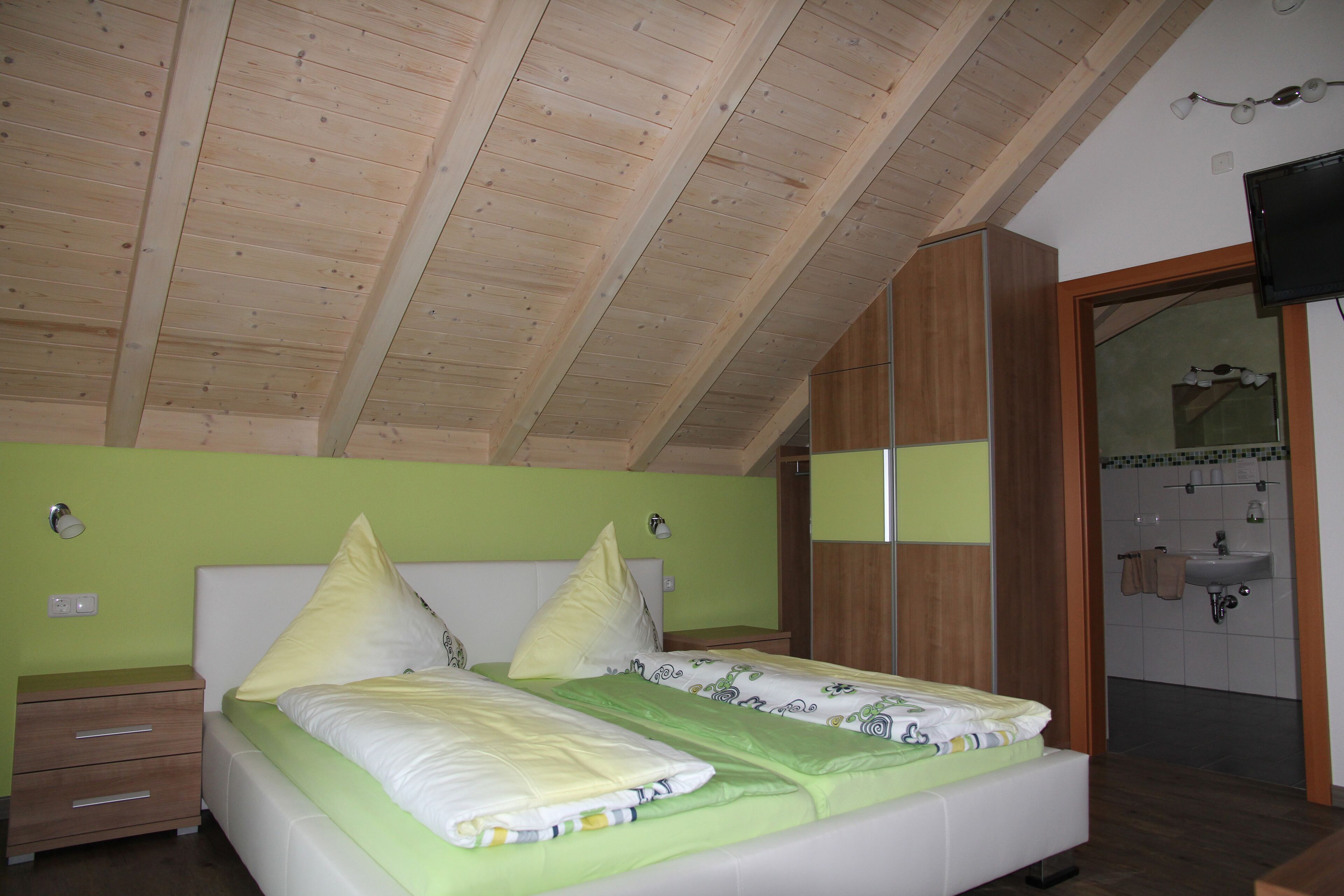 Un dormitorio con techo de madera, una cama con ropa de cama verde, una mesita de noche y un baño visible a través de una puerta abierta.