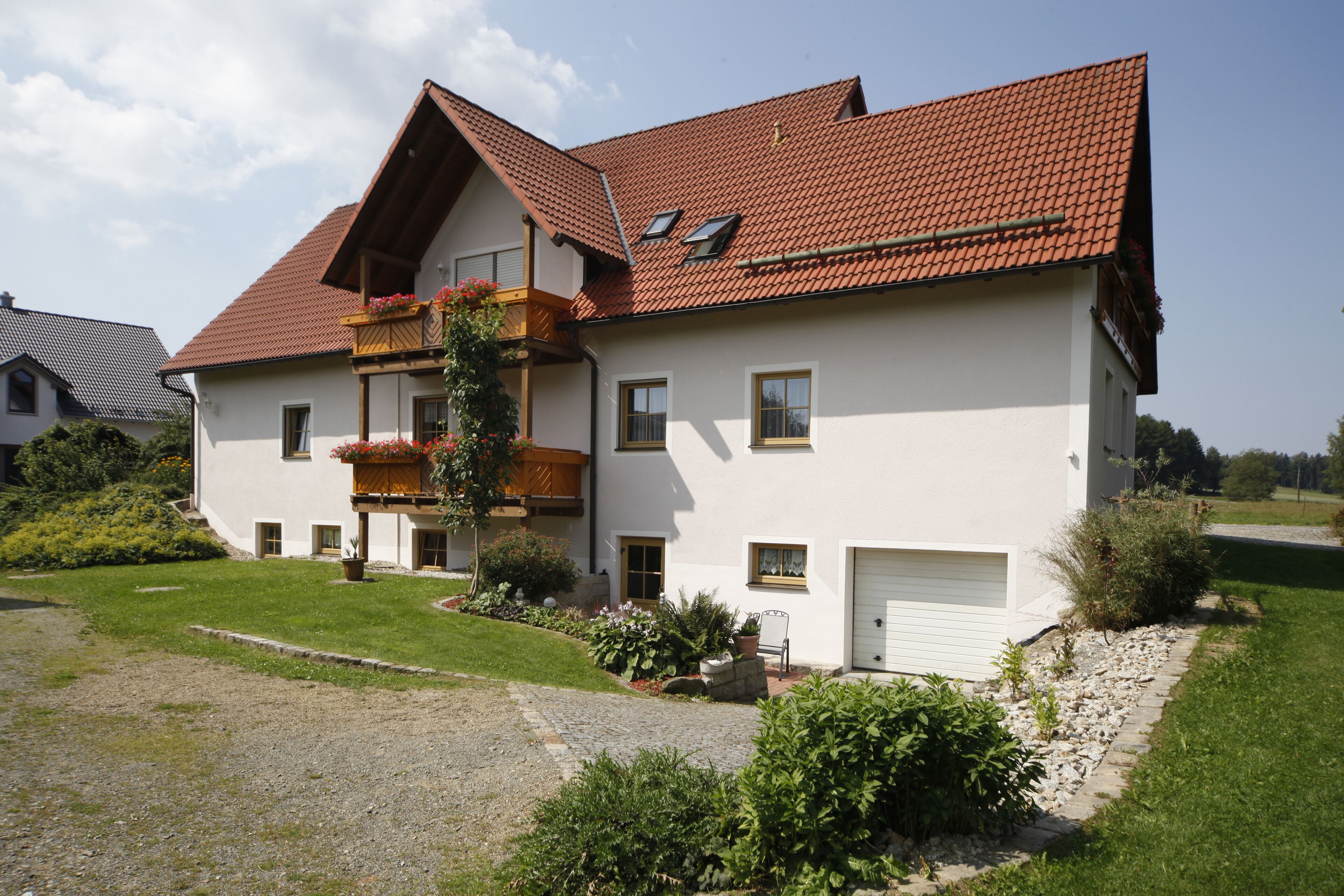A two-story house with a sloped roof, a balcony with plants, and a gravel driveway.