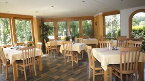 A restaurant with multiple tables and chairs, large windows, and a view of greenery outside.