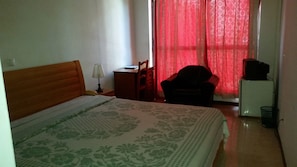 A bedroom with a bed, a chair, a desk, a lamp, and a refrigerator. - Hotel América (Praia)