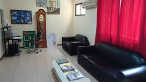A living room with a black sofa, a white coffee table with magazines, a wooden clock, and a red curtain. - Hotel América (Praia)