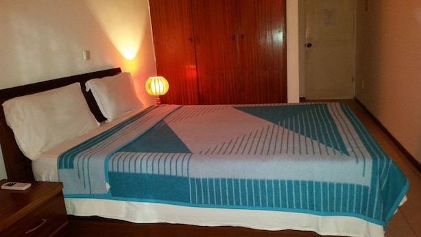 A neatly made bed with a striped blanket, a bedside table with a remote control, and a wall-mounted lamp. - Hotel América (Praia)