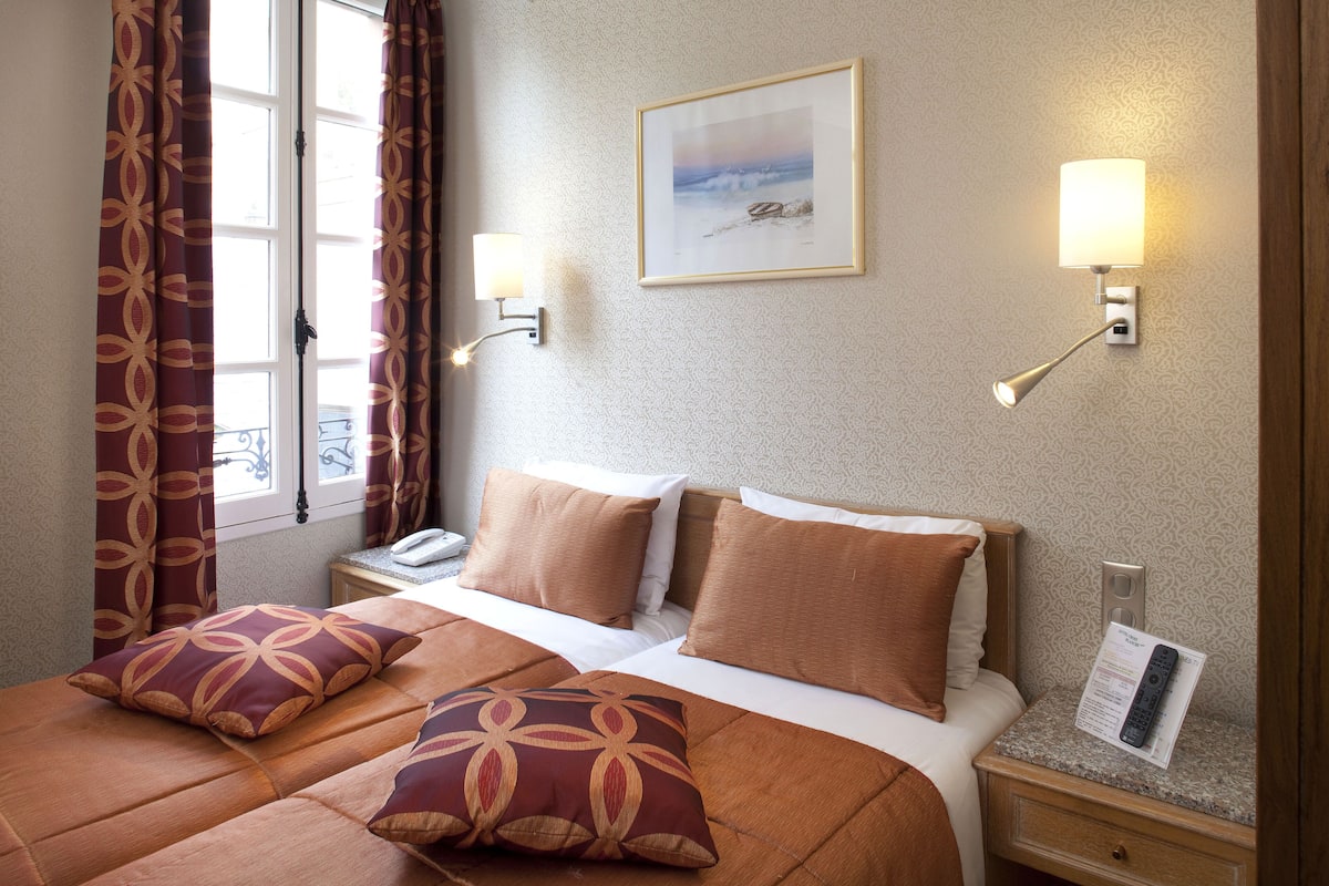 Habitación de hotel con una cama, dos almohadas, una mesita de noche, una lámpara, un cuadro enmarco y una ventana con cortinas.