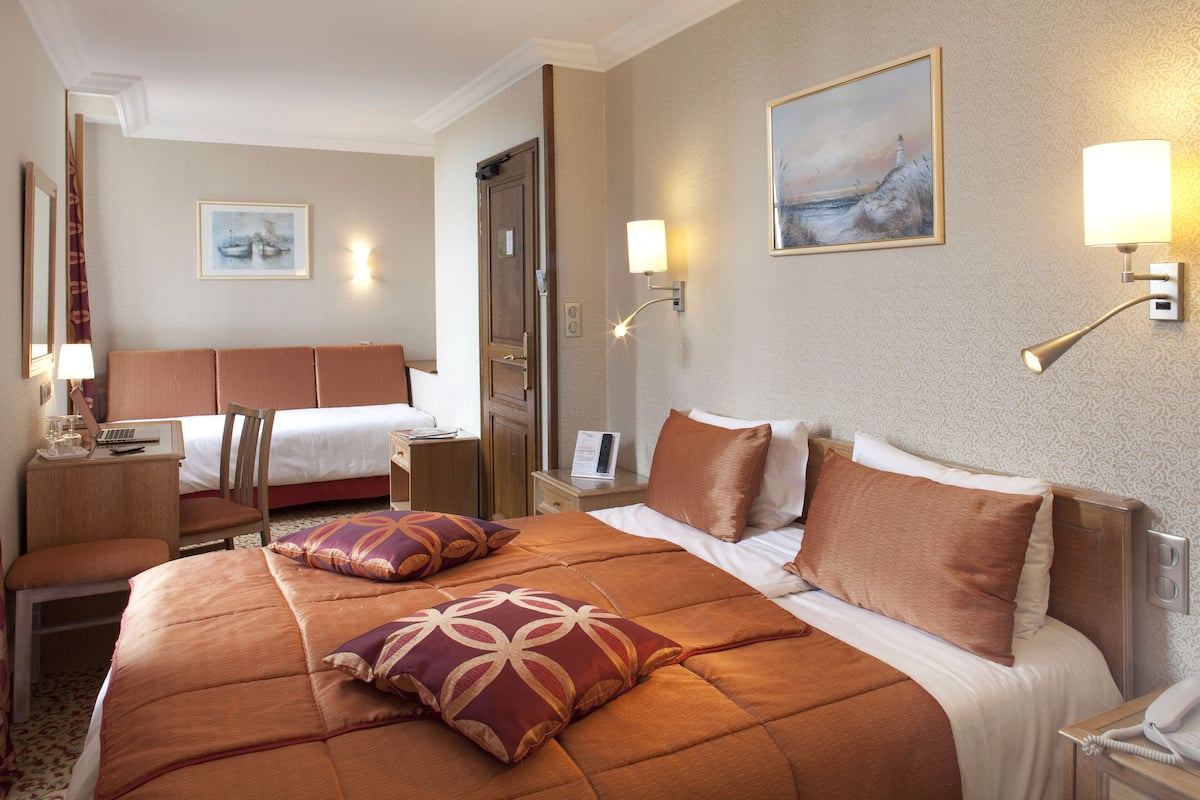 Habitación de hotel con una cama grande, una cama más pequeña, un escritorio con lámpara, una silla y un cuadro en la pared.