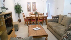 A living room with a wooden coffee table, a sofa, a dining table with chairs, and a bookshelf. - Ahr237 - Hamlets at West Haven - 4 Bed 3 Baths Villa (Davenport)