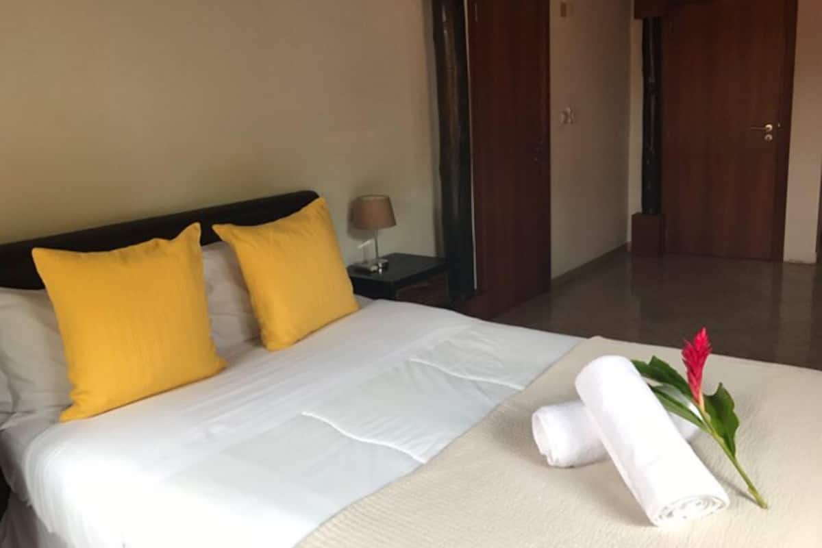 Une chambre d’hôtel avec un lit, deux oreillers jaunes, un couvre-lit blanc, une table de chevet avec une lampe et une serviette roulée ornée d’une fleur rouge, posée sur le lit.