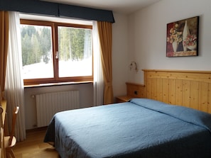 A bedroom with a bed, a window with curtains, a radiator, a wooden headboard, a small table, and a painting on the wall.