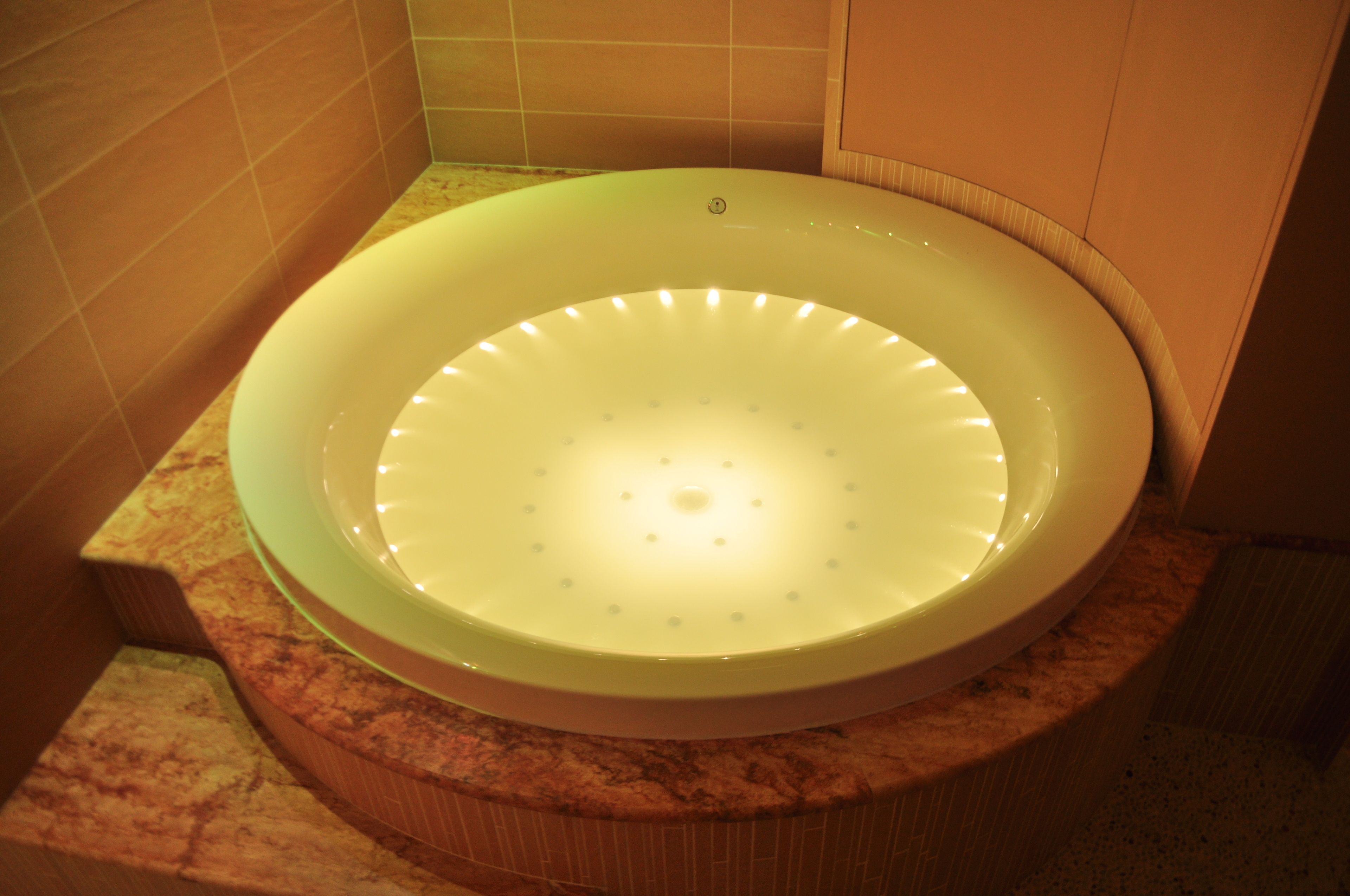 Una bañera circular con una lámpara en un baño con paredes de azulejos y encimera de mármol.
