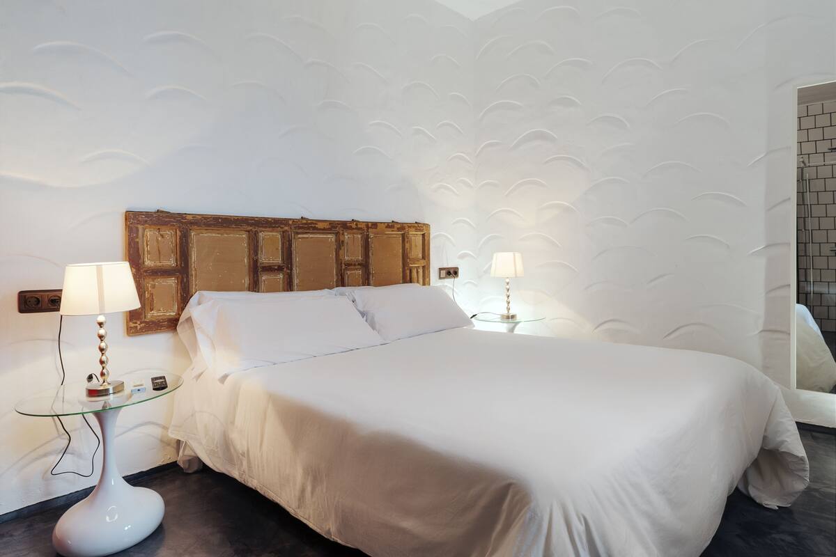 Un lit bien fait, avec du linge de lit blanc, deux tables de chevet avec des lampes et un mur texturé derrière le lit.