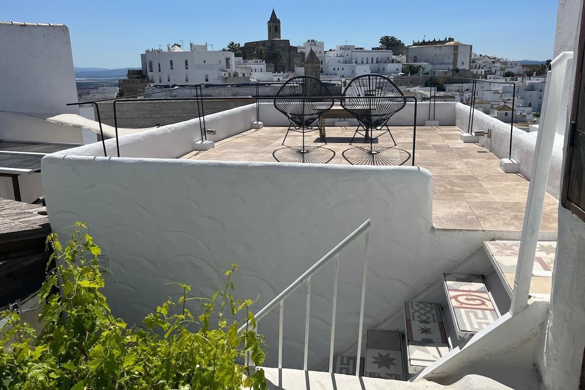Une terrasse sur le toit offrant une vue sur une ville historique, avec un bâtiment blanc doté d’un clocher et deux chaises en métal noir.