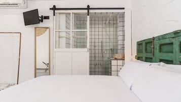 Un dormitorio con cama, puerta corredera hacia el baño, televisor montado y una puerta verde.