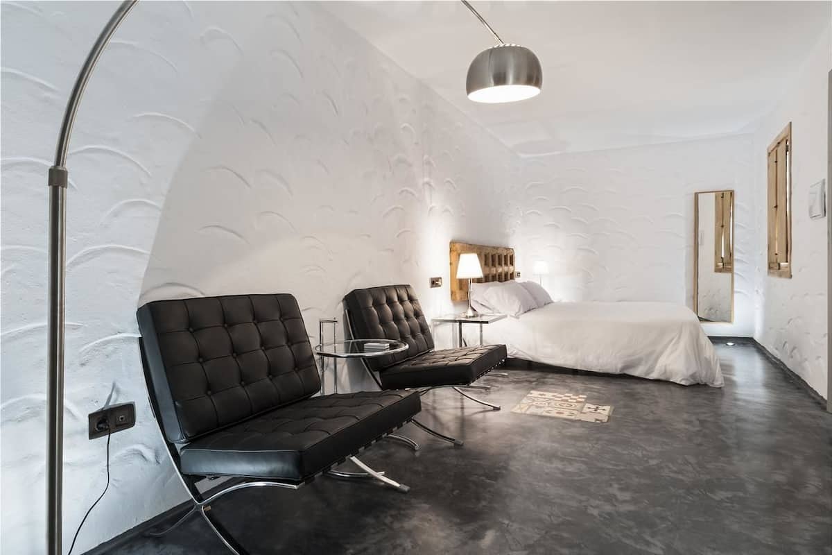 Une chambre à coucher moderne avec un lit, deux chaises noires, une petite table ronde, une lampe sur pied et un miroir au mur.
