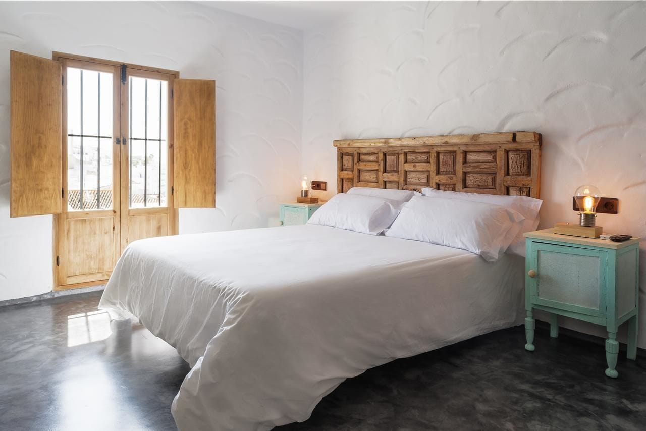 Un dormitorio con cabecera de madera, cama con ropa de cama blanca, dos mesitas de noche con lámparas y un ventanal con contraventanas de madera.