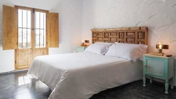 Un dormitorio con cabecera de madera, cama con ropa de cama blanca, dos mesitas de noche con lámparas y un ventanal con contraventanas de madera.