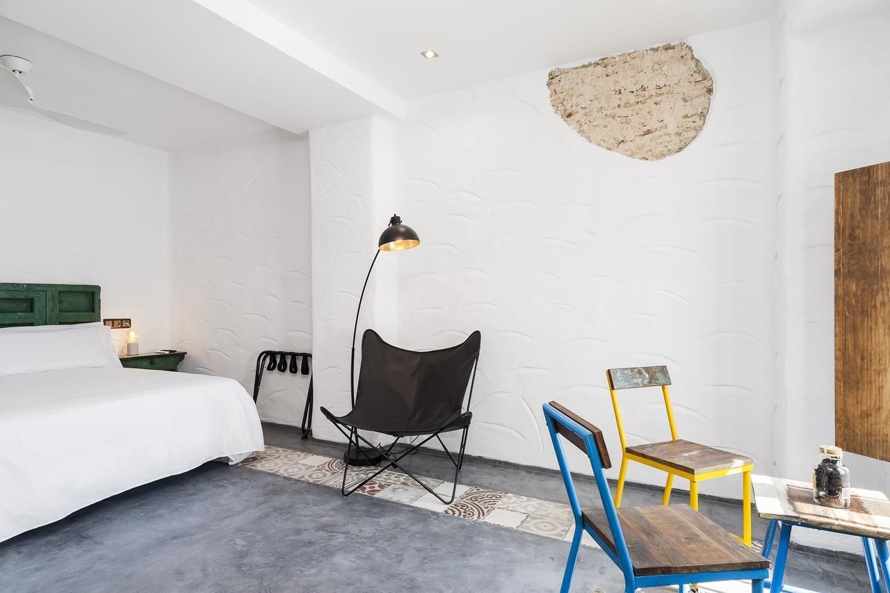 A hotel room with a bed, a black folding chair, a yellow and blue chair, a small table with a bottle, and a unique textured wall.