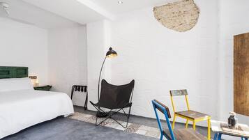 Habitación de hotel con cama, una silla plegable negra, una silla amarilla y azul, una mesita con una botella y una pared de textura única.