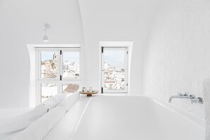 Ein modernes Badezimmer mit einer großen Badewanne, zwei Fenstern mit Blick auf die Stadt und einem kleinen runden Tisch mit Kerzen.