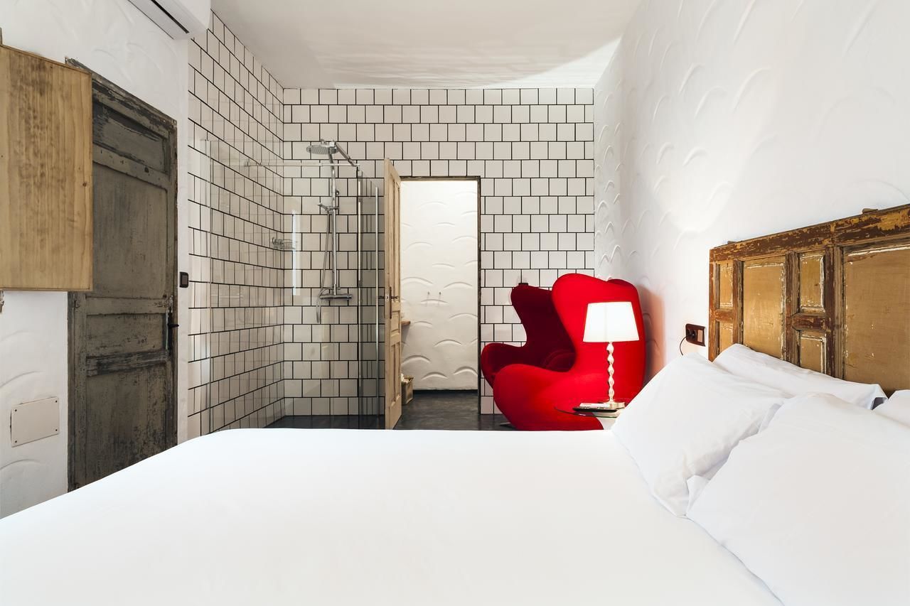Quarto de hotel com uma cama, uma cadeira vermelha, chuveiro e porta de madeira.