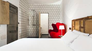 Quarto de hotel com uma cama, uma cadeira vermelha, chuveiro e porta de madeira.