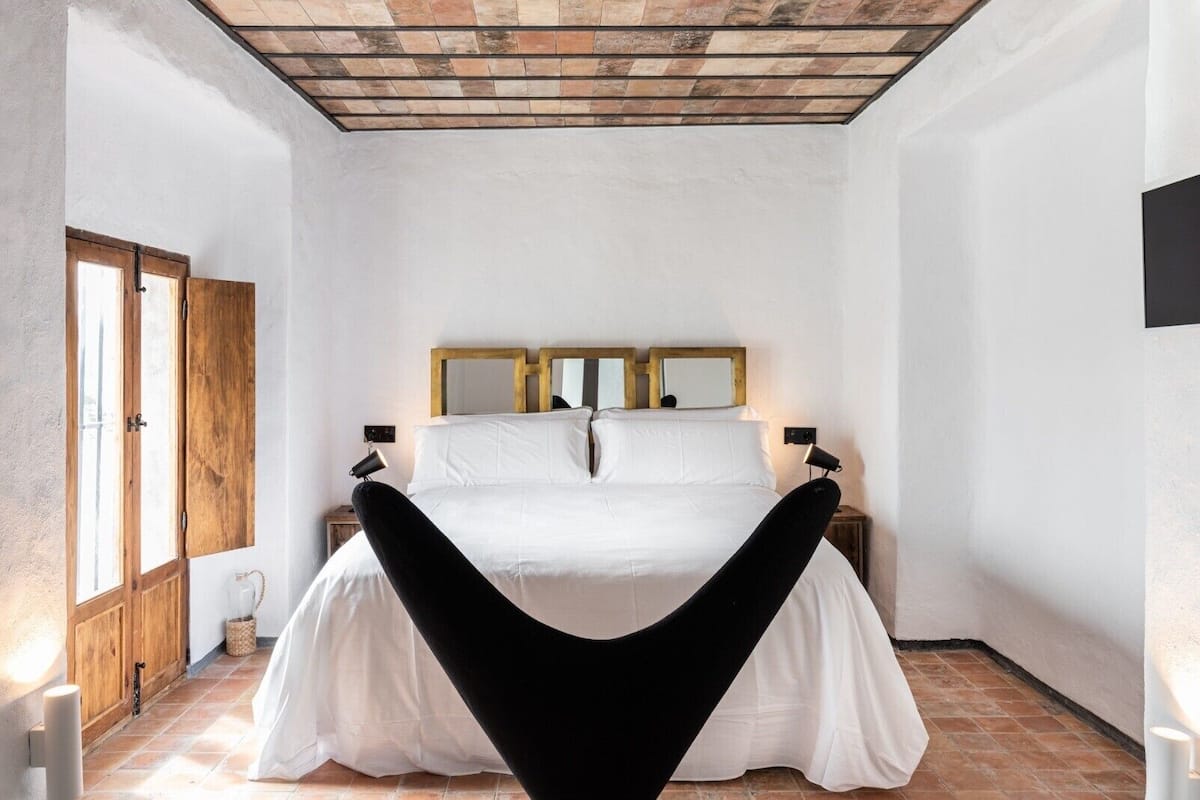 Une chambre à coucher avec un lit placé au centre, sous un plafond en pente, et flanqué d’armoires en bois et d’un grand miroir.