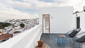 Une terrasse sur le toit offrant une vue sur une ville aux maisons blanchies à la chaux, une porte portant l’inscription « 10 », et deux chaises avec des coussins.
