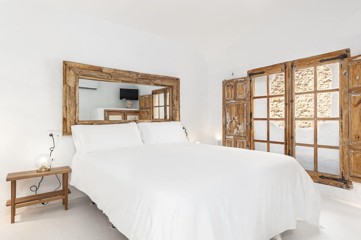 Une chambre à coucher avec un grand lit, une table de chevet en bois avec une lampe, un miroir et une fenêtre avec un cadre en bois.