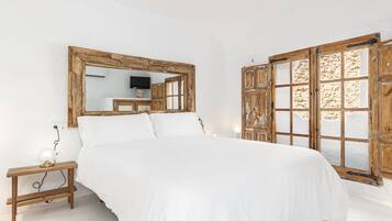 A bedroom with a large bed, a wooden nightstand with a lamp, a mirror, and a window with a wooden frame.