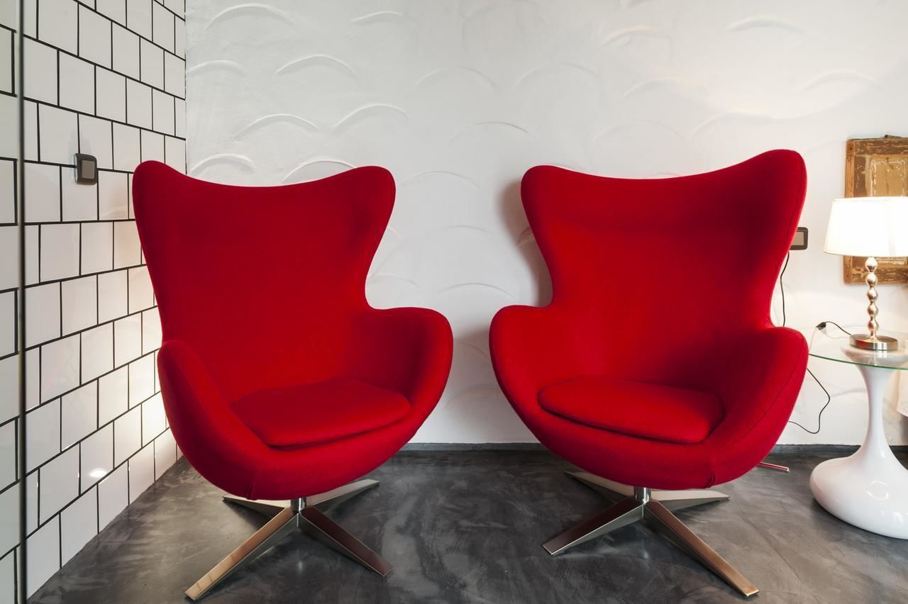 Dos sillas rojas con patas metálicas, una pared blanca con un diseño de olas y una mesa blanca con una lámpara.