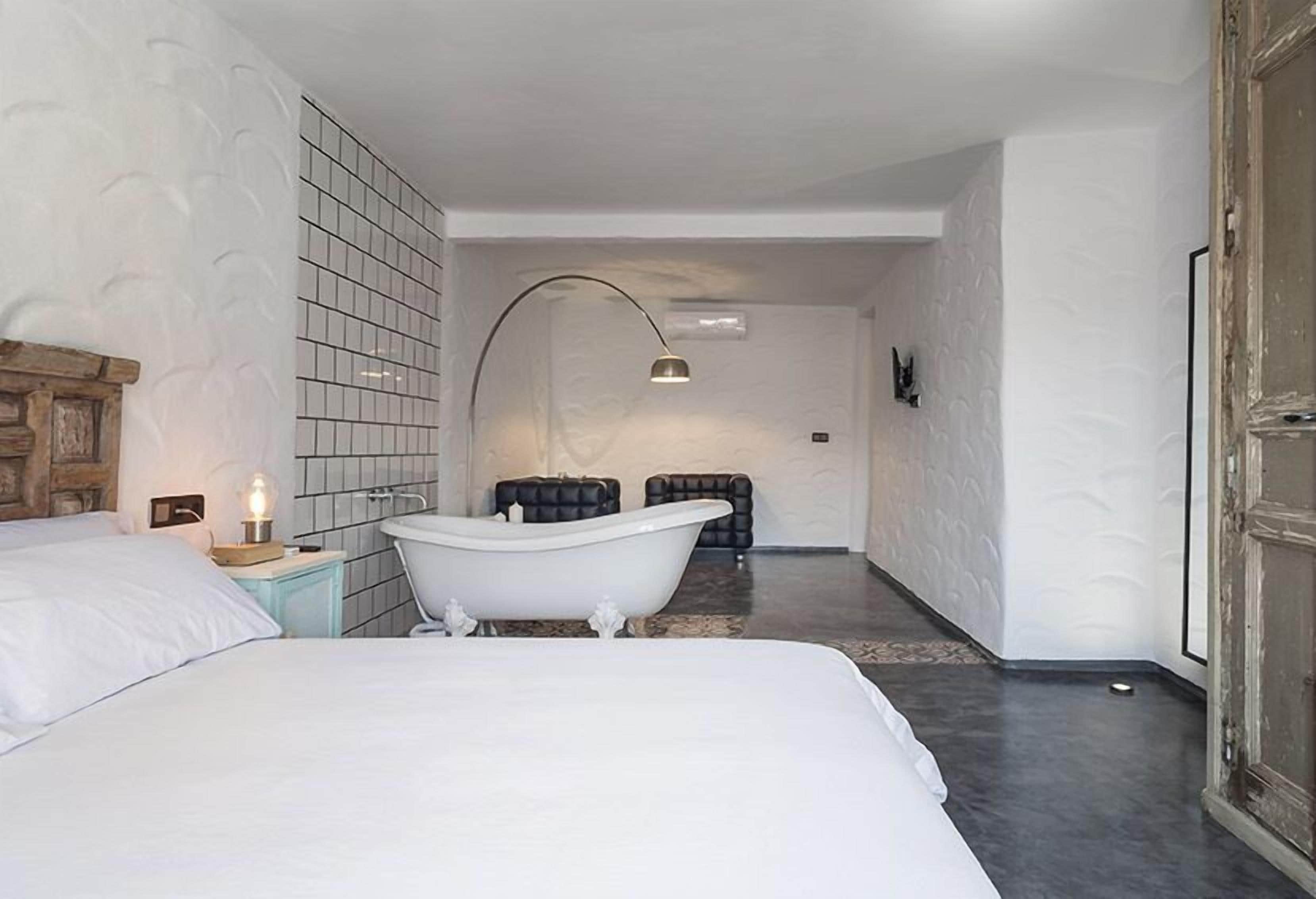 A modern hotel room with a large bed, a white bathtub, a small bedside table with a lamp, and a large mirror.