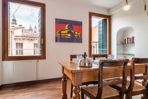 Zona de menjador amb taula i cadires de fusta, un radiador i vista a edificis des de la finestra.
