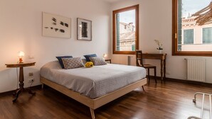 Une chambre à coucher avec un lit en bois, une petite table, une chaise et une fenêtre donnant sur des immeubles.