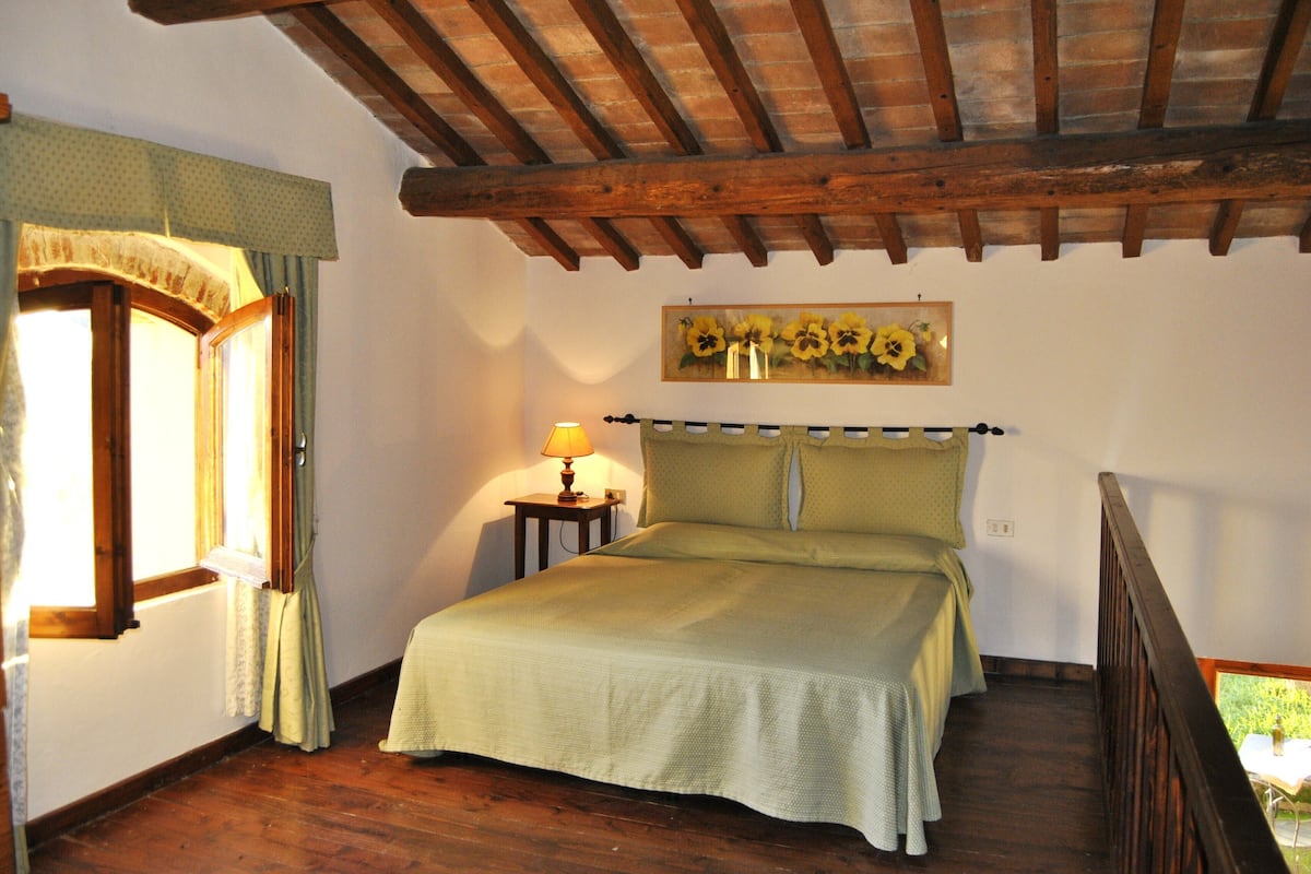 Een slaapkamer met een houten plafond, een bed met groene beddengoed, een nachtkastje met een lamp en een raam met gordijnen.