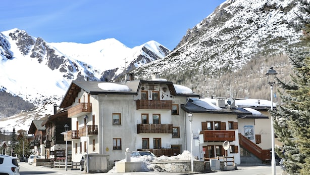 A mountain lodge with multiple balconies, a stone facade, and a view of snow-covered mountains. - Appartamenti Brunnlein (Livigno)