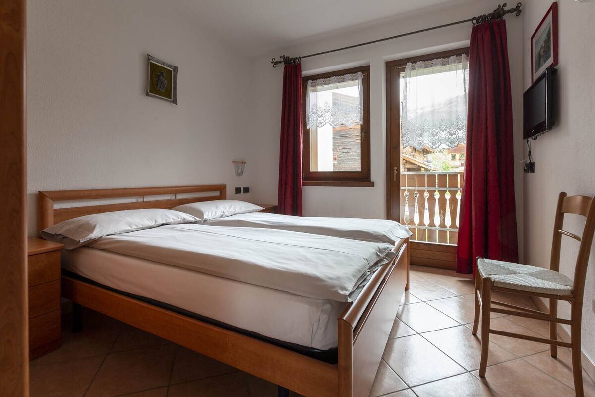 A double bed with white linens, a wooden nightstand, a chair, a small table, and a window with red curtains.