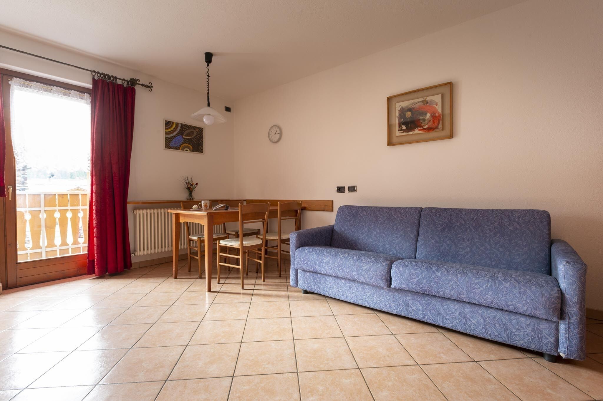 Un salon avec un canapé bleu, une table à manger avec des chaises, un radiateur et une fenêtre avec des rideaux rouges.