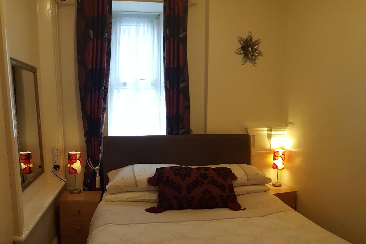 A hotel room with a bed, two bedside lamps, a nightstand, a mirror, and a window with curtains.